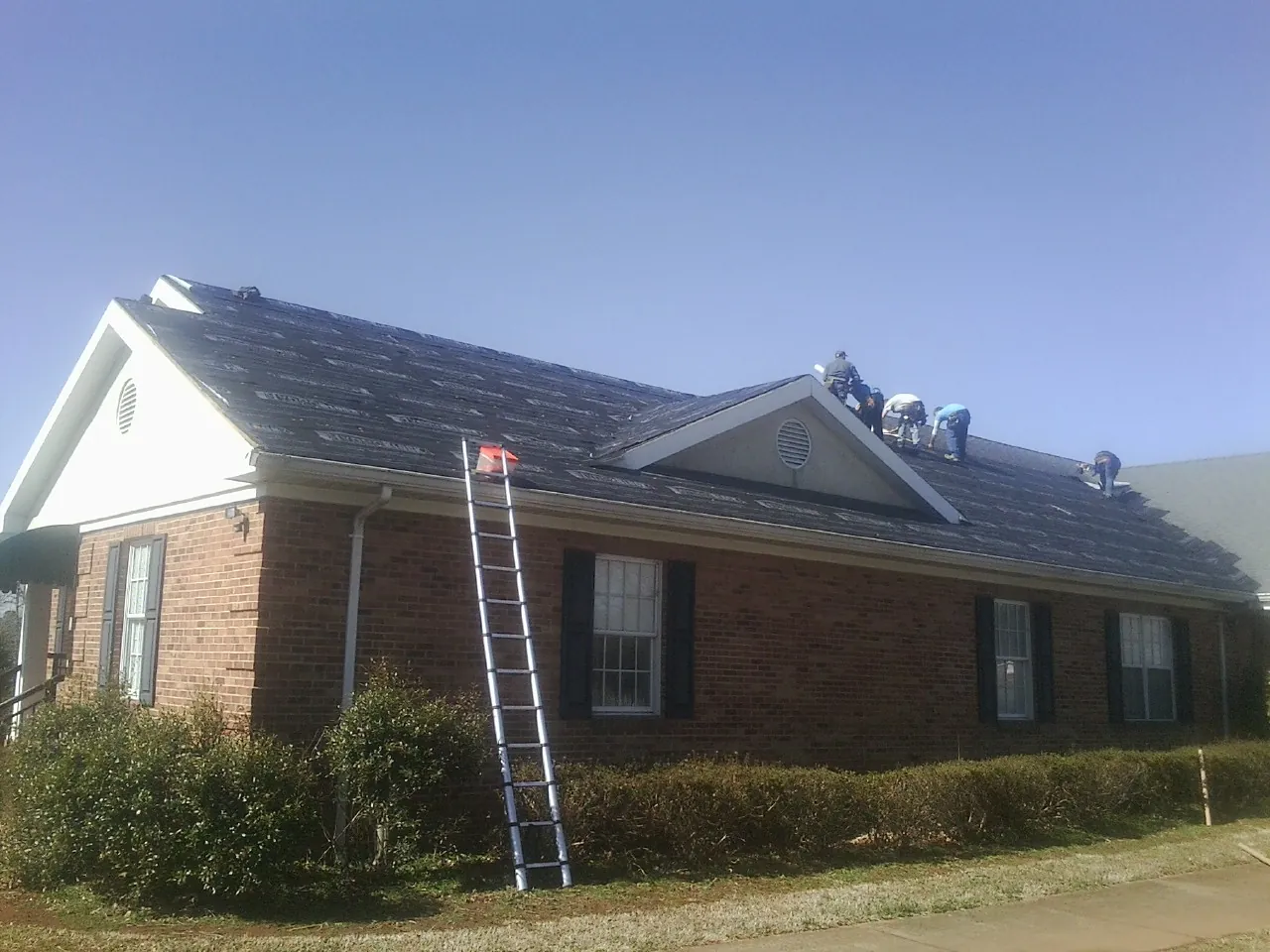 Crew tearing shingles off a brick ranch roof with a ladder set — Emergency Roof Repair work by our crew in Park Falls