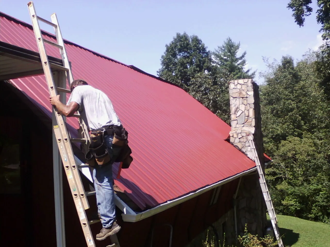 Roofer on a ladder beside a red metal roof and stone chimney — Soffit & Fascia Repair by Roofing Brothers in Park Falls